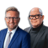 Zwei Männer, einer lächelnd im blauen Anzug mit Brille, der andere im schwarzen Rollkragenpullover mit schwarzer Brille.