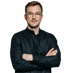 Ein junger Mann mit Brille und schwarzem Hemd mit verschränkten Armen vor grünem Hintergrund.