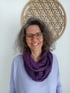 Lächelnde Frau mit Brille, grau meliertem Haar, lila Schal und Wandschmuck Blume des Lebens im Hintergrund.