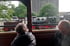 Zwei Personen blicken von einem Waggon auf eine grüne Dampflokomotive mit rotem Fahrgestell.