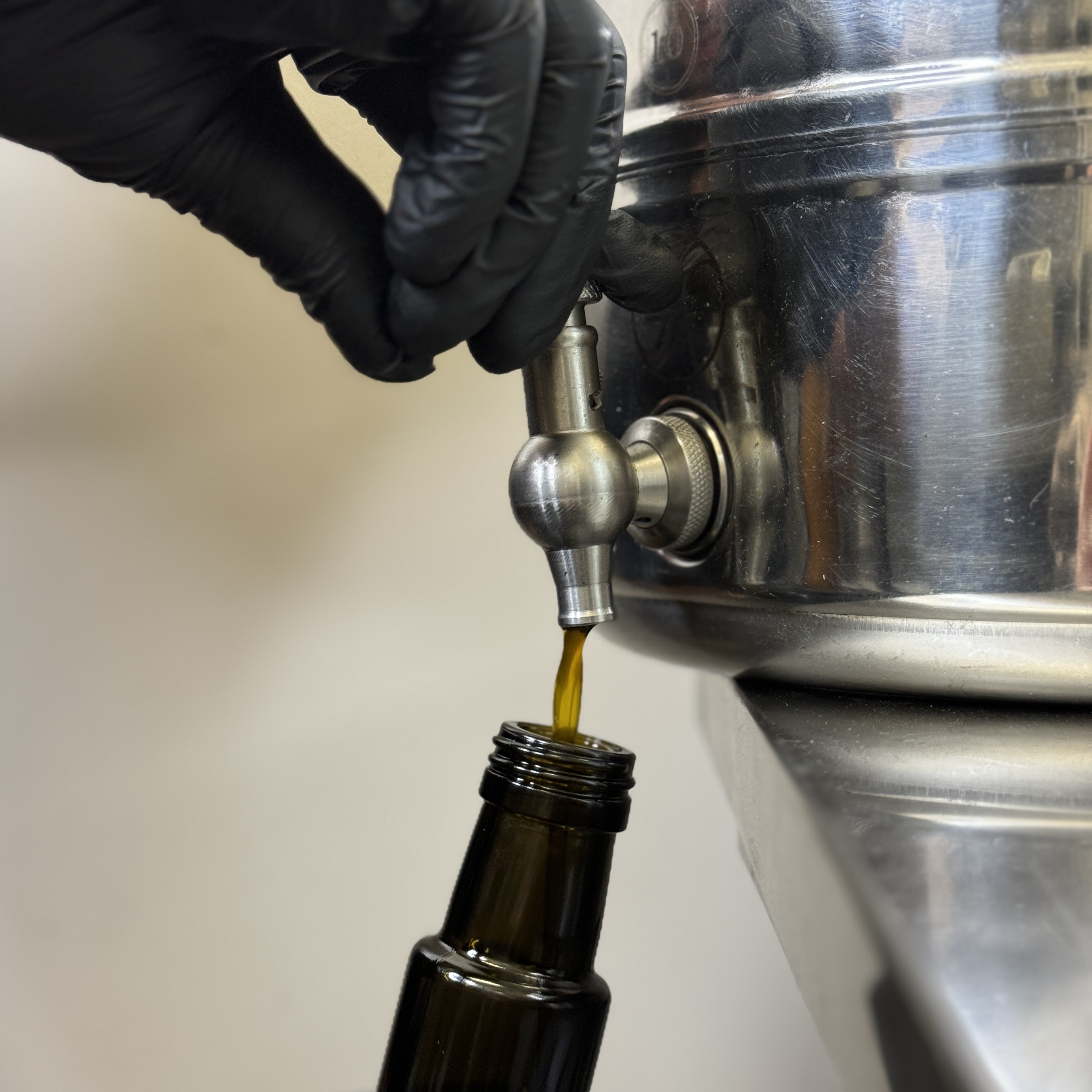 Schwarzer Handschuh öffnet Edelstahlventil am Sedimentationsbehälter — goldgelbes Öl läuft in dunkle UV-Glasflasche