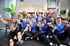 Lachende Gruppe von 14 Personen in blauen T-Shirts, die ihre Arme heben, vor der Aufschrift „jd performance“.