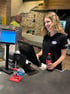 Lächelnde Frau in schwarzem Esso-Polohemd an einer Ladenkasse, hält eine Coca-Cola-Flasche. Ein Mann steht im Hintergrund.