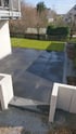 Moderne Terrasse aus großen, dunkelgrauen Steinplatten grenzt an eine grüne Rasenfläche mit Holzzaun im Hintergrund.