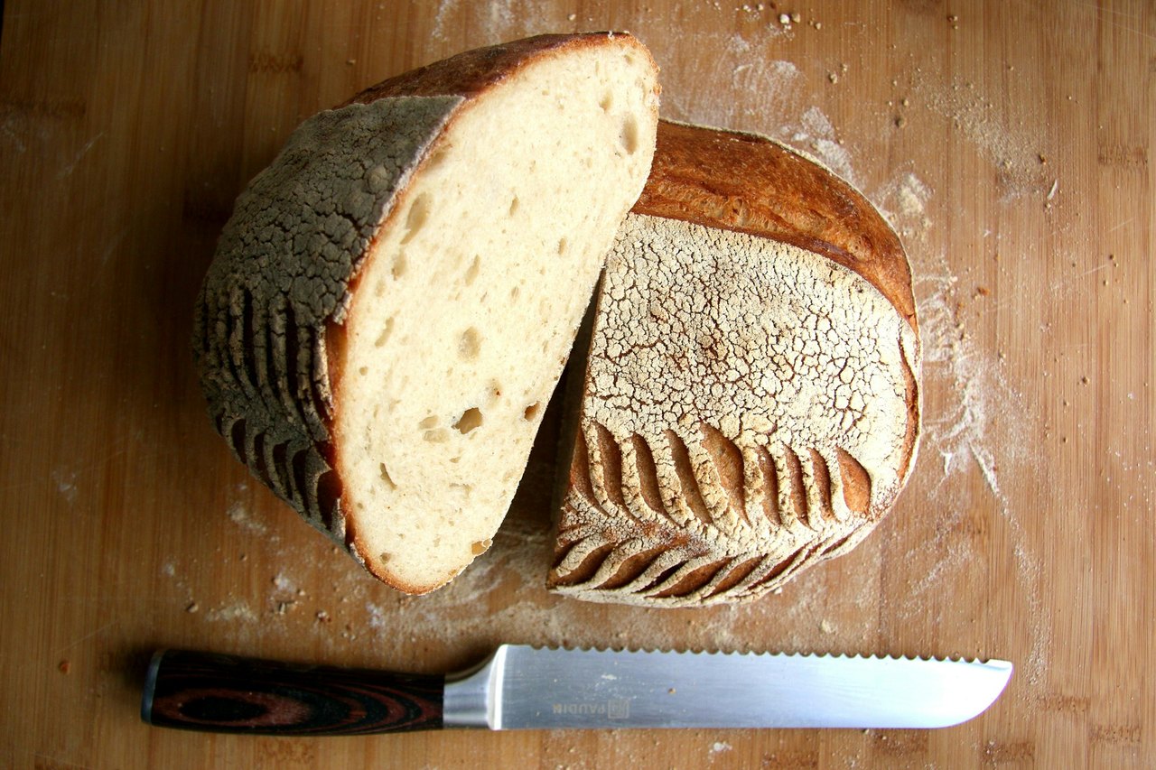 Ein angeschnittener Brotlaib liegt auf einem bemehlten Holzbrett, davor ein Brotmesser, ringsum Krümel und Mehlstaub.