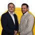 Zwei lächelnde Männer in Anzügen stehen vor einem gelben, abstrakten Design.