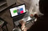 Person sitzt mit Teetasse vor einem Laptop, auf dessen Bildschirm eine Website mit bunten Kacheln und Text zu sehen ist.