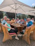 Eine Gruppe Senioren sitzt an einem Holztisch in einem Biergarten unter weißen Veltins-Sonnenschirmen mit Getränken.