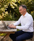 Ein lächelnder Mann im weißen Hemd arbeitet mit einem Laptop an einem Holztisch im grünen Garten.