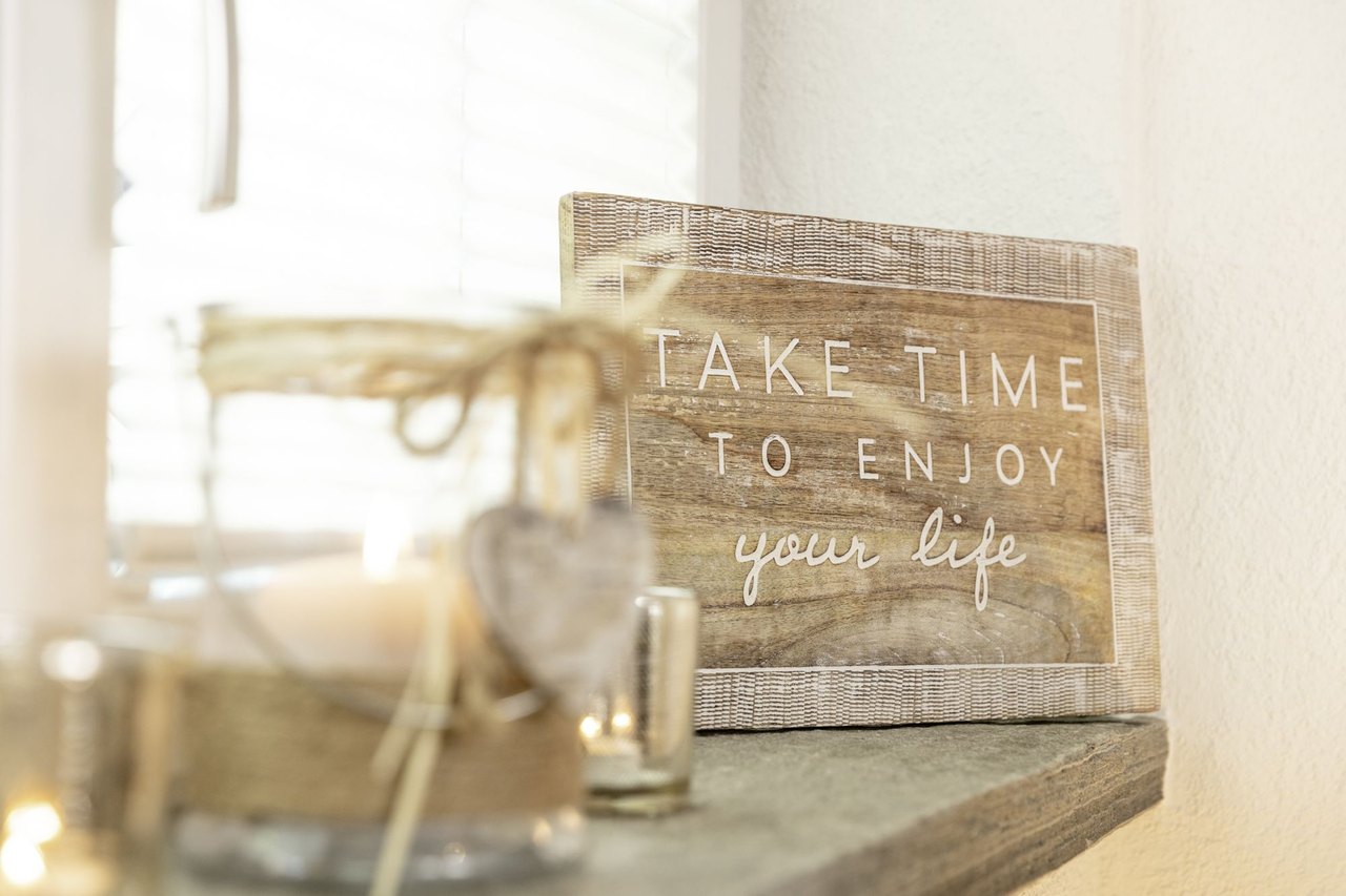 Holzschild mit der Aufschrift Take time to enjoy your life auf dem Fenstersims der Praxis, davor Kerzen im Gegenlicht