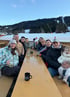 Eine Gruppe von Menschen sitzt lächelnd an einem langen Holztisch im Freien vor einer verschneiten Bergkulisse.