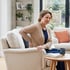 A woman sits in a beige armchair holding her lower back in discomfort while looking away.