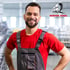 Ein lächelnder Mann in grauer Latzhose und rotem T-Shirt steht in einer Fabrikhalle; Werner Zodel Logo.