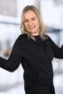 Eine lächelnde Frau mit blonden Haaren und schwarzer Kapuzenjacke steht vor einem unscharfen Hintergrund.