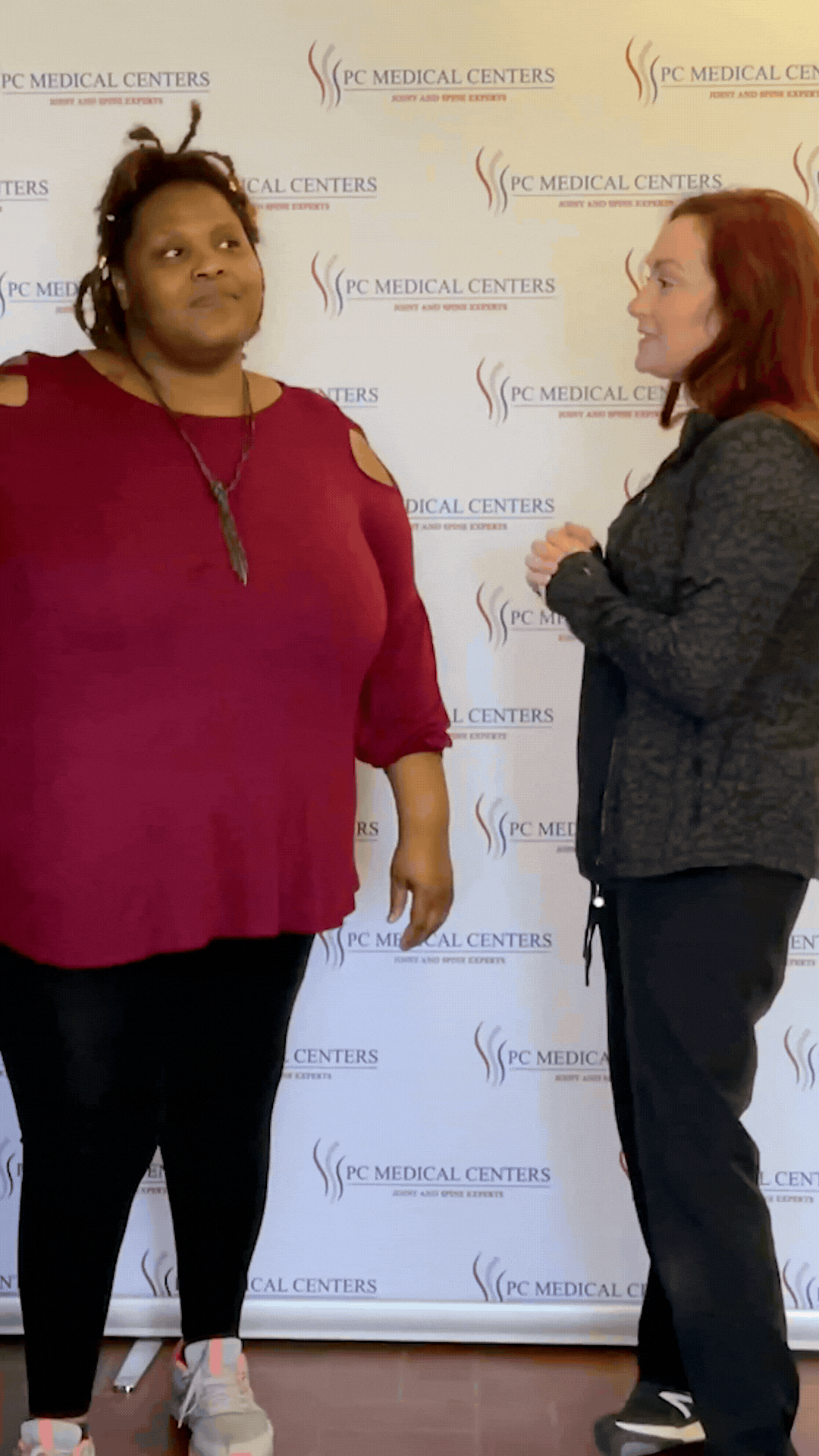 Patient in a red top talking with a PC Medical Centers staff member in front of the clinic's branded backdrop