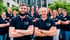 Lächelndes Team von 17 Personen in dunklen Poloshirts mit gekreuzten Armen, draußen vor einem Gebäude.