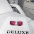Weiße Schutzbrille mit rosa Gläsern auf einem weißen Handtuch mit schwarzem "DELUXE" Logo auf einer Behandlungsliege.