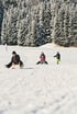 Drei Kinder rodeln lachend einen verschneiten Hügel hinunter, umgeben von schneebedeckten Tannen.