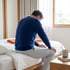 A man in a blue sweater sits on the edge of a bed with his head bowed and hands on his knees.