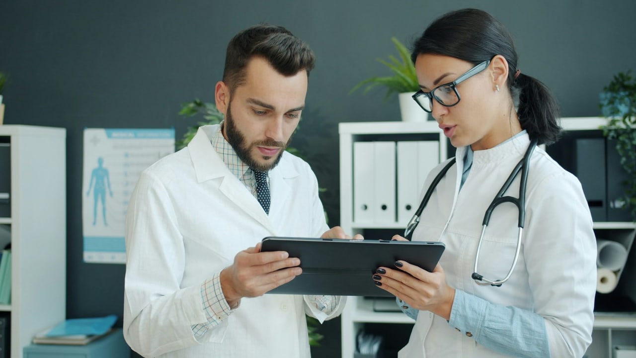 Arzt und Ärztin im Kittel besprechen etwas auf einem Tablet im Sprechzimmer