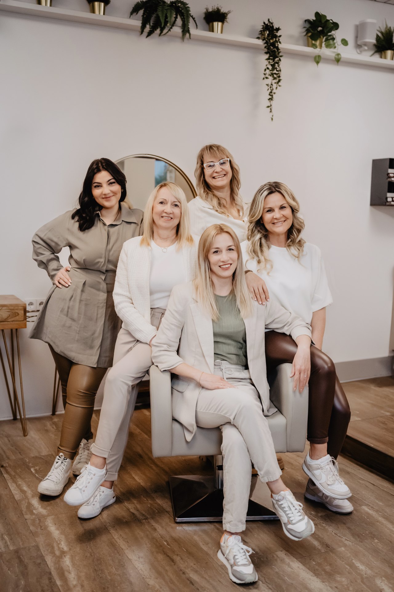 Fünf lächelnde Frauen in einem hellen Friseursalon posieren gemeinsam um einen Friseurstuhl.