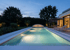 Beleuchteter Swimmingpool mit weißer Einfassung, dahinter eine Terrasse und ein Haus, bei Dämmerung.