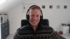 Ein lächelnder Mann mit Headset und gemustertem Strickpullover sitzt in einem hellen Büro vor einer Webcam.