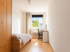 Helles schmales Zimmer mit Holzbett, Schreibtisch am Fenster, hellem Laminatboden und weißem Kleiderschrank.