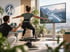 Frau macht Kniebeugen auf einem Balance-Board vor einem Bildschirm mit virtueller Berglandschaft, betreut von einem Trainer.
