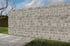 Eine graue Mauer aus gestapelten Betonsteinen, dahinter grüner Rasen und ein Baum.