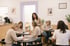 Eine Lehrerin im beigen Blazer steht beratend neben Frauen, die an runden Tischen in einem hellen Seminarraum schreiben.