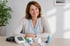 Lächelnde Frau mit Blutdruckmessgerät, Medikamentenblistern und Asthma-Inhalator auf dem Tisch.