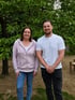 Eine lächelnde Frau im rosa Hoodie und ein Mann im weißen T-Shirt stehen nebeneinander vor einem grünen Laubbaum im Freien.