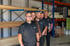 Das Singer und Singer Team in schwarzen Poloshirts stehen vor Holzregalen in einem Lager.