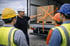 Ein Mann im Schutzhelm zeigt auf eine mit orangefarbenen Gurten gesicherte Palette mit Kartons auf der Ladefläche eines Lkw.