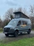 Mercedes Sprinter Reisewagen mit geöffnetem Aufstelldach auf Kopfsteinpflaster – individueller Van-Ausbau im modernen Grau vor bewaldetem Hintergrund