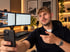 Mann mit Bart macht ein Selfie im Home-Office mit zwei Monitoren, Tastatur und Mikrofon vor einer beleuchteten Holzwand.