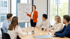 Frau in roter Jacke zeigt auf ein Flipchart mit Diagramm vor einer Gruppe von fünf Personen am Tisch.