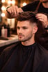 Friseur schneidet die Haare eines jungen Mannes mit Bart, der einen schwarzen Umhang trägt.