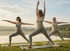 Drei Frauen unterschiedlichen Alters machen Yoga in Kriegerpositionen auf Matten am Seeufer.