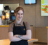 Lachende junge Frau mit gefalteten Armen, McDonald's Schürze und Tattoo, an einer Theke stehend.