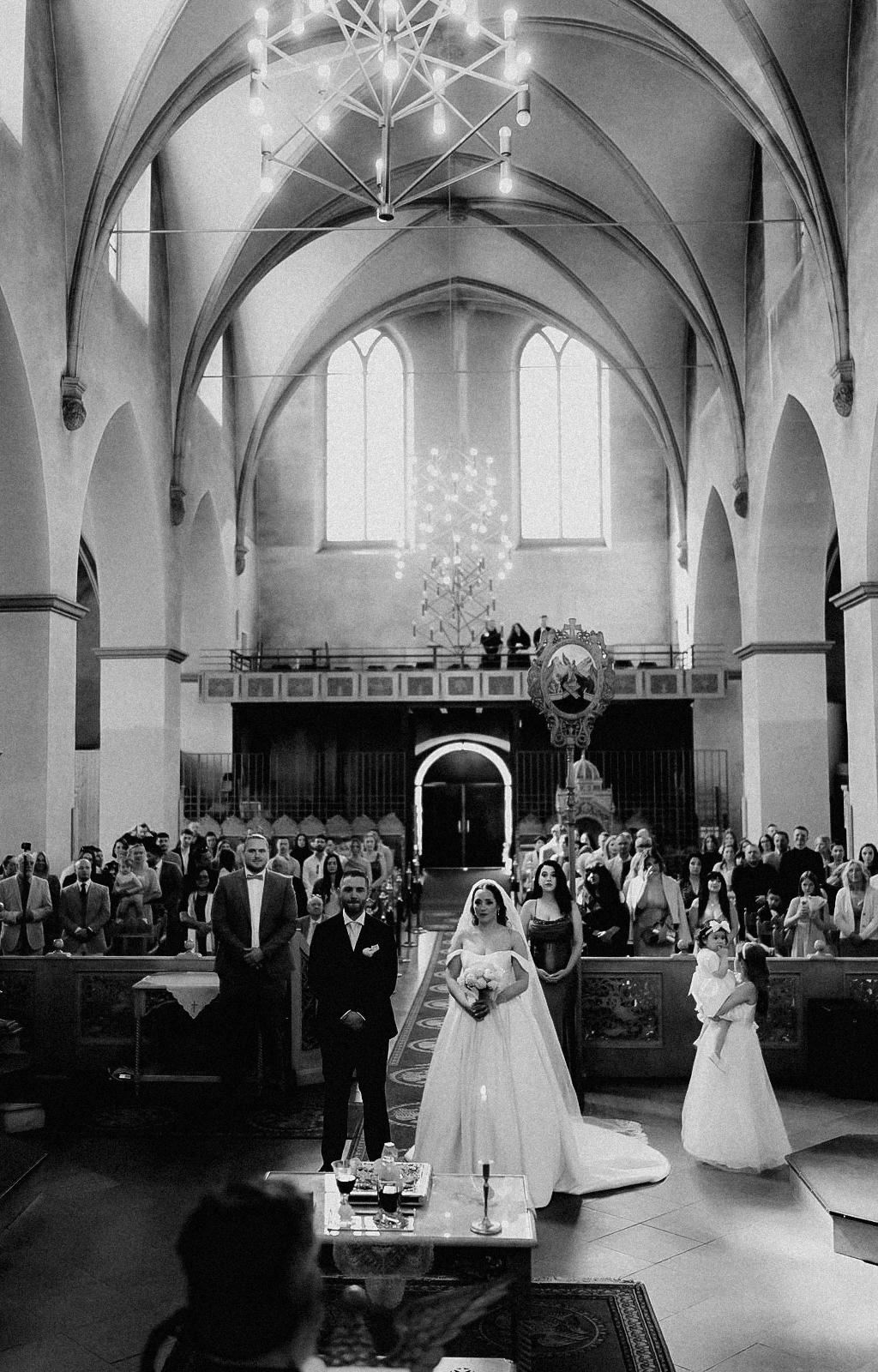 Schwarz-Weiß-Aufnahme: Weiter Blick durch die orthodoxe Kirche in Köln während der Trauung, Brautpaar und Blumenkind stehen vorn im Mittelgang, die Gäste in den Bankreihen unter dem hohen Gewölbe.