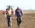 Zwei Bauern gehen über ein gepflügtes Feld; ein Traktor ist im Hintergrund.