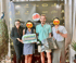 Four people, two women and two men, holding gifts in front of a dark banner with event logos and "TOP GROUP."