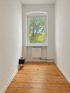 Leerer, schmaler Raum mit Dielenboden, weißem Heizkörper unter dem Fenster und Blick ins Grüne.