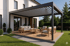 Moderne Pergola mit Lamellendach über einer Holzterrasse mit Lounge-Möbeln vor einem weißen Haus.