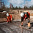 Zwei Bauarbeiter in orangefarbener Warnkleidung und weißen Helmen verlegen Bewehrungsstahl auf einer Baustelle im Freien.