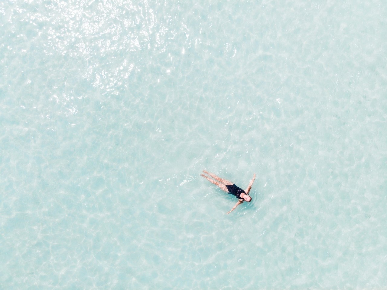 Ein Mensch treibt mit ausgebreiteten Armen rücklings auf hellem, ruhigem Wasser, von weit oben fotografiert.