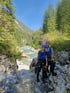 Zwei Personen in Neoprenanzügen und Helmen stehen an einem felsigen Flussbett in einer Berglandschaft.