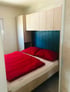 Bett mit roter Bettwäsche, blauem Kopfteil und hellen Holzschränken darüber in einem kleinen Raum.